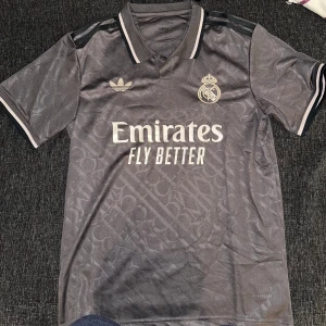 Grå Real Madrid fotbollströja - Snygg grå Real Madrid fotbollströja från Adidas med korta ärmar och klassisk krage. Tröjan har klubbens emblem och Adidas-logga i vitt samt texten 'Emirates Fly Better' på framsidan. Perfekt för alla fotbollsfans!