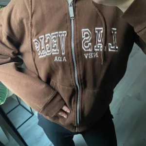 Brun hoodie med Las Vegas-tryck - Säljer en brun hoodie med dragkedja och Las Vegas-tryck på framsidan. Den har en mysig huva och långa ärmar.