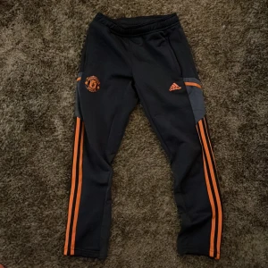 Manchester united träningsbyxor från Adidas - Snygga marinblåa träningsbyxor från Adidas med orangea detaljer och Manchester United-logga.