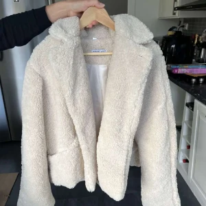 Beige teddyjacka  - Mysig beige teddyjacka med fluffig textur och krage. Perfekt för kyliga dagar när du vill hålla dig varm och stilren. Jackan har en klassisk passform och är lätt att matcha med olika outfits.