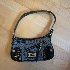 Mönstrad axelväska från Gussaci - Snygg axelväska från Gussaci med ett unikt mönster i svart och grått. Väskan har en dragkedja och dekorativa detaljer i svart lack. Perfekt för att ge din outfit en extra touch.