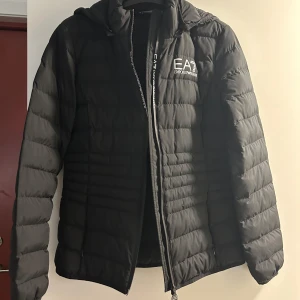 Svart pufferjacka från Emporio Armani - Snygg svart pufferjacka unisex från Emporio Armani EA7 med dragkedja och huva. Perfekt för kyliga dagar med sin quiltade design som håller värmen. Jackan har en stilren look med logotypdetalj på bröstet. Storlek M. Finns i Stockholm. 