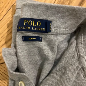 Grå pikétröja från Ralph Lauren - Snygg grå pikétröja från Ralph Lauren i slim fit. Tröjan har en klassisk krage och knappar framtill. Den är dekorerad med det ikoniska broderade logotypen i mörkblått på bröstet.