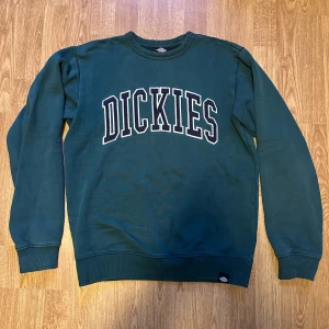 Grön sweatshirt från Dickies - Säljer en grön sweatshirt från Dickies med stort tryck på framsidan. Tröjan har långa ärmar och ribbade muddar vid ärmslut och nederkant. Liten defekt vid trycket. Går att klippa bort, men lägger priset på denna med det i åtanke!