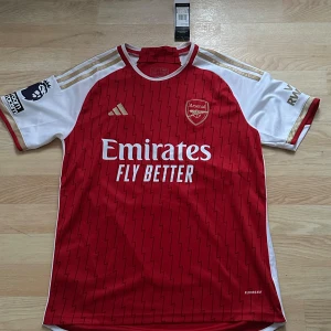 Röd Arsenal fotbollströja från Adidas - Snygg röd Arsenal fotbollströja från Adidas med vita och guldiga detaljer. Har aldrig använt. Storlek är M/S