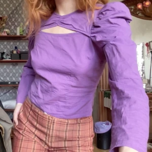 Lila topp med puffärmar - Snygg lila topp med puffärmar och unik cut-out detalj vid halsen. Toppen har en åtsittande passform.  Längd: 49cm Axel till axel: 54cm Ärm: 68cm