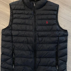 Svart dunväst från Ralph Lauren - Snygg svart dunväst från Ralph Lauren med dragkedja och broderad logga i rött på bröstet. Perfekt för lager-på-lager under kyliga dagar.