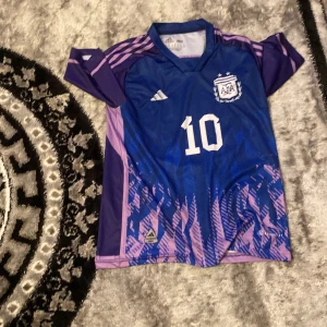 Blå och lila fotbollströja med nummer 10 - Snygg blå och lila fotbollströja från Adidas med nummer 10 och namn på ryggen. Tröjan har korta ärmar och ett coolt mönster i lila och rosa nyanser. Perfekt för fotbollsfans som vill sticka ut på planen! Storlek S
