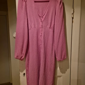 Rosa klänning från Sisters Point - Charmig rosa klänning från Sisters Point med v-ringning och knappar framtill. Klänningen har långa ärmar och en texturerad yta som ger en unik look. Perfekt för både vardag och speciella tillfällen.