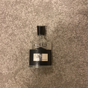 Creed Aventus parfym - Säljer en exklusiv Creed Aventus parfym i en elegant glasflaska med svart etikett och silverdetaljer. Flaskan har en stilren design med en svart kork. Perfekt för den som vill ha en lyxig doftupplevelse.