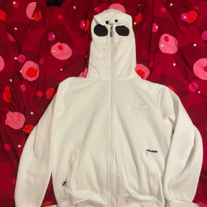 Vit Fullzip hoodie från C.P. Company - Säljer en vit huvtröja från C.P. Company med unika glasögon i huvan. Tröjan har dragkedja framtill och två fickor med dragkedjor. Perfekt för en stilren och modern look.