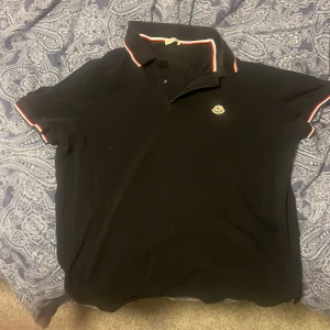 Svart kortärmad pikétröja från Moncler - Säljer en stilren svart pikétröja från Moncler med korta ärmar. Tröjan har en klassisk krage med röd och vit randig detalj samt Monclers logotyp broderad på bröstet. Perfekt för en avslappnad men ändå elegant look.