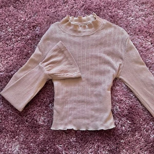 Beige ribbad tröja med volangdetaljer - Beige ribbad tröja med volangdetaljer vid halsen och ärmsluten. Tröjan har långa ärmar och en croppad passform i stretchigt material. Strl XS.