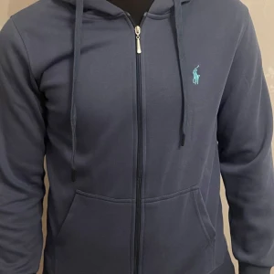 Mörkblå hoodie från Ralph Lauren - Snygg mörkblå hoodie från Ralph Lauren med dragkedja och huva. Den har en broderad logga på bröstet och långa ärmar. Perfekt för en avslappnad stil. Jag säljer den då den är väldigt liten på mig, köpte den för ett tag sen och har slutat använda den, inga skador alls.