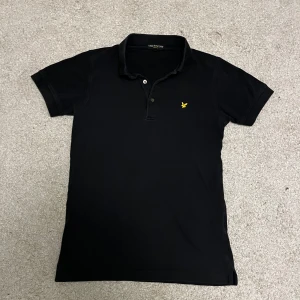 Svart pikétröja från Lyle & Scott - Snygg svart pikétröja från Lyle & Scott med klassisk krage och knappar. Den har en klassiska gula logotypen på bröstet. Perfekt för sommaren eller under en half zip mm. Bra skick och använd sparsamt, inga defekter. Storlek S.