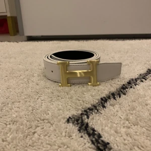 Vitt bälte med guldfärgat spänne - Snyggt vitt bälte med ett stort guldfärgat H-spänne. Perfekt för att ge en stilren touch till din outfit. Bältet är smalt och elegant, vilket gör det lätt att matcha med olika kläder.