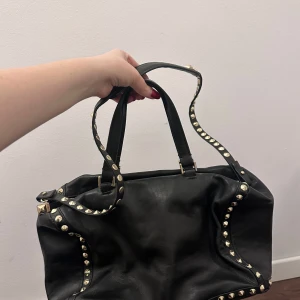 Svart axelväska med nitar - Snygg svart axelväska i skinn med coola nitar längs kanterna. Väskan har en praktisk dragkedja upptill och dubbla handtag för enkel bärning. Perfekt för att ge din outfit en edgy touch.