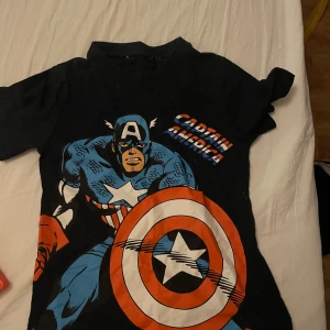Svart Captain America t-shirt - Cool svart t-shirt med Captain America-tryck i blått, rött och vitt. Perfekt för superhjältefans! 👊