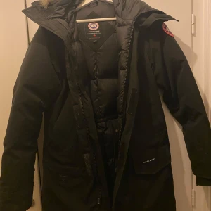 Svart parkajacka från Canada Goose - Säljer en svart parkajacka från Canada Goose med pälskantad huva. Jackan har en lång design med dragkedja och knappar framtill. Perfekt för kalla vinterdagar med sin varma dunfyllning. Ikonisk logga på ärmen. Sliten på armarna längst ner men syns ej när man har den på sig 