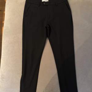 Snygga svarta kostymbyxor från Les Deux i slim fit. Byxorna har en klassisk design med knappar och dragkedja framtill. Perfekta för en stilren look.