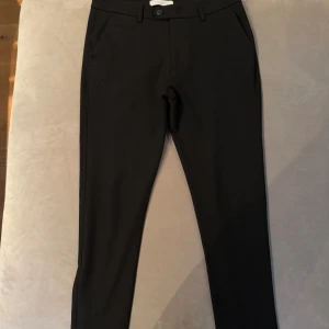 Svarta kostymbyxor från Les Deux - Snygga svarta kostymbyxor från Les Deux i slim fit. Byxorna har en klassisk design med knappar och dragkedja framtill. Perfekta för en stilren look.