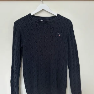 Svart kabelstickad tröja från Gant - Snygg svart kabelstickad tröja från Gant med rund halsringning och broderad logotyp på bröstet. Perfekt för kyligare dagar och ger en stilren look.