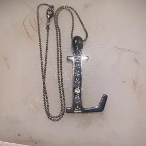 Silverfärgat halsband med bokstaven L - Snyggt silverfärgat halsband med ett hängsmycke i form av bokstaven L, dekorerat med glittrande stenar. Perfekt för att ge en personlig touch till din stil. OBS EN STEN HAR RAMLAT AV!
