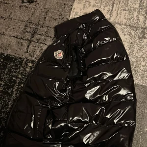 Svart pufferjacka från Moncler - Säljer en svart, glansig pufferjacka från Moncler. Jackan har en quiltad design och en dragkedja framtill. Perfekt för kyligare väder och ger en stilren look.