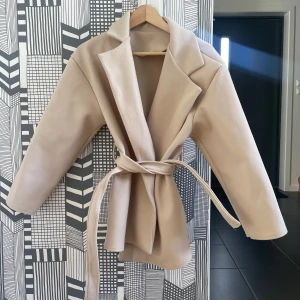 Beige kort kapp - Beige super fin kort kappa😍 kappan har två rymliga fickor och ett band runt midjan. Den är i nyskick o är helt oanvänd. Perfekt nu till hösten😍