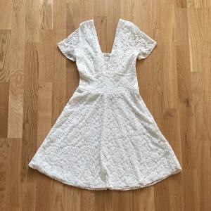 Vit spetsklänning - Säljer en superfin vit klänning i spetsmaterial. Den har en v-ringad design och korta ärmar, perfekt för sommarens alla fester. Klänningen är i nyskick och har en normal passform som ger en elegant look. Den är både bekväm och stilren, vilket gör den till ett måste i garderoben!