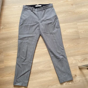 Grå kostymbyxor från Carin Wester - Säljer ett par stilrena grå kostymbyxor från Carin Wester. De har en klassisk design med dragkedja och knapp framtill samt hällor för bälte. Byxorna har en rak passform och är perfekta för både kontoret och mer formella tillfällen.