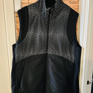 Svart väst från SOC - Säljer en svart väst från SOC i storlek L. Västen har ett coolt mönster upptill med vita detaljer och en dragkedja framtill. Perfekt för höst och vår när du vill hålla dig varm men ändå se stilren ut. Materialet känns slitstarkt och bekvämt.