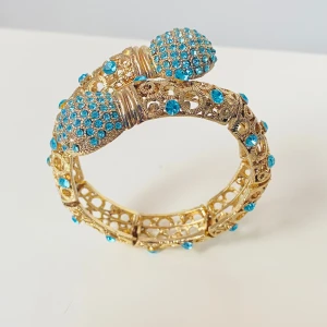 Guldarmband med turkosa stenar - Säljer ett supercoolt guldarmband med massor av turkosa stenar. Armbandet har en unik design med två stora dekorationer som är täckta av små turkosa stenar. Armbandet är i bra skick, saknar någon enstaka rhinestone men syns knappt 