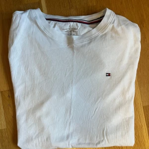 Vit t-shirt från Tommy Hilfiger - Säljer en klassisk vit t-shirt från Tommy Hilfiger med en liten broderad logotyp på bröstet. T-shirten har en rund halsringning med röd och blå randig detalj på insidan av kragen.