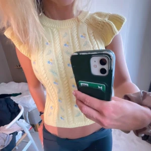 Stickad topp  - Säljer en söt gul stickad topp med korta volangärmar och ett fint blommönster i blått och vitt. Toppen är croppad och har en ribbad struktur som ger en snygg passform. Perfekt för vår och sommar!