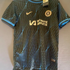 Svart och blå Chelsea fotbollströja - Snygg Chelsea fotbollströja från Nike i svart med blått rutmönster. Tröjan har korta ärmar och klubbens logga på bröstet. Perfekt för fans som vill visa sitt stöd. Shorts medföljer 