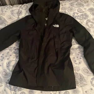 Svart jacka från The North Face - Säljer en svart jacka från The North Face med huva och dragkedja. Jackan har ett stilrent utseende och är perfekt för utomhusaktiviteter. Den är fodrad med ett mönstrat innerfoder för extra komfort. PASSAR SOM XS