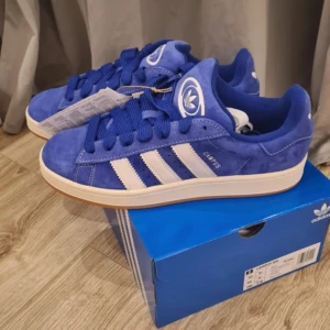 Blå Adidas Campus sneakers - Snygga blå Adidas Campus sneakers i mocka med vita ränder och detaljer. Skorna har en klassisk design med snörning och en gummisula i kontrasterande färg. Perfekta för en stilren och sportig look.