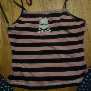 Randig linne med dödskallemotiv - Säljer ett randigt linne i svart och rosa med smala axelband och spetsdetalj vid halsen. Linnet har ett coolt dödskallemotiv med en liten rosett på framsidan. Perfekt för en avslappnad stil.