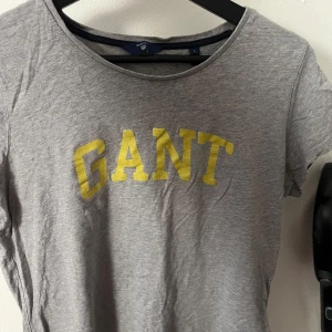 Grå t-shirt från GANT - Säljer en grå t-shirt från GANT med gult tryck på framsidan. Köpt från Zalando, den har endast en liten liten fläck som syns på tredje bilden annars ingenting. Strl L, den har krympt ganska mycket så passar mer som en S nu 