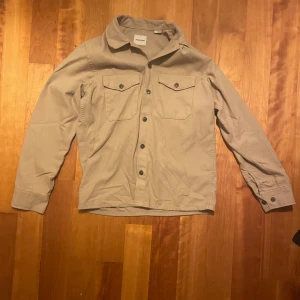 Beige overshirt från Jack & Jones - Snygg beige overshirt med långa ärmar och knappar framtill. Två praktiska bröstfickor med knappar ger en stilren look. Perfekt för lager-på-lager under höst och vår.