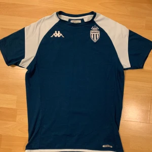 Ac Monaco träningströja kappa blå - Snygg blå fotbollströja från Kappa med AS Monaco FC-emblem på bröstet. Tröjan har korta ärmar och vita detaljer på axlarna. Köpt i Monaco för runt 500kr, inga problem alls. Storlek s men passar m