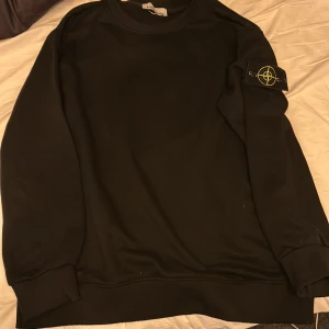 Svart sweatshirt från Stone Island - Säljer en stilren svart sweatshirt från Stone Island med deras ikoniska märke på ärmen. Tröjan har långa ärmar och ribbade muddar vid ärmslut och nederkant. Perfekt för en casual look.