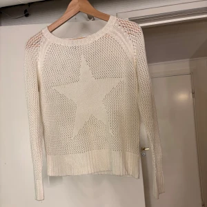 Vit stickad tröja med stjärna - Säljer en vit stickad tröja med en stor stjärna i mitten. Tröjan har långärmade och ett luftigt stickat mönster som ger en avslappnad look. Perfekt för lager-på-lager under kyliga dagar.