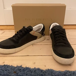 Burberry skor - Snygga svarta sneakers från Burberry med klassiskt rutigt mönster på sidorna. Skorna har en vit sula och snörning framtill. Perfekta för en stilren look.