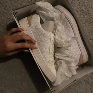 Vita sneakers från Michael Kors - Snygga vita sneakers från Michael Kors med diskret logotypmönster och snörning. Perfekta för en stilren look.
