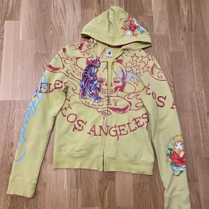 Gul hoodie med tryck Ed hardy y2k vtg 90 size M  - Säljer en unik gul hoodie med färgglada tryck av en tiger och texten 'Los Angeles'. Hoodien har en dragkedja framtill och en stor huva. Perfekt för dig som vill sticka ut med en cool och färgglad stil.