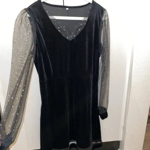 Svart klänning med glittriga ärmar - Elegant svart klänning i sammet med långa, transparenta ärmar dekorerade med glittriga detaljer. Klänningen har en v-ringad hals och en stilren design som passar perfekt för festliga tillfällen.