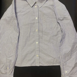 Blå och vit randig skjorta från H&M - Snygg blå och vit randig skjorta från H&M med klassisk krage och knappar framtill. Perfekt för en stilren look.