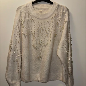 Vit stickad tröja med pärlor - Säljer en elegant vit stickad tröja dekorerad med pärlor på framsidan och ärmarna. Tröjan har en rund halsringning och långa ärmar, perfekt för att ge en stilren look. Passar utmärkt för kyligare dagar.